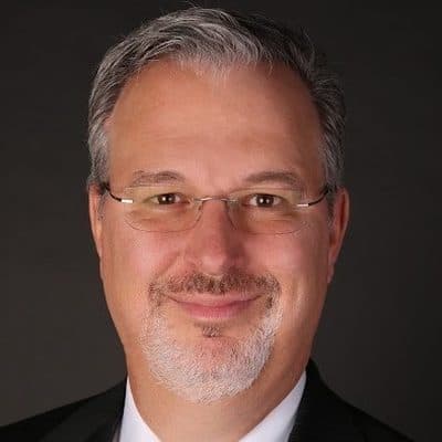 Gregory S. DuPont, in a dark suit and wire-rimmed glasses, against a dark studio backdrop