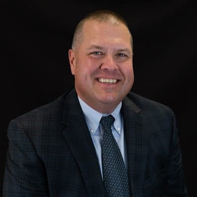 Mike Moeller, in a checked navy jacket and tie, against a dark studio backdrop