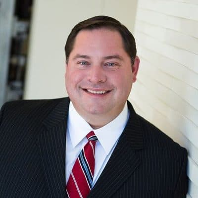Reagan Allen, in a dark pinstripe suit and red striped tie, smiling in front of a pale stone wall